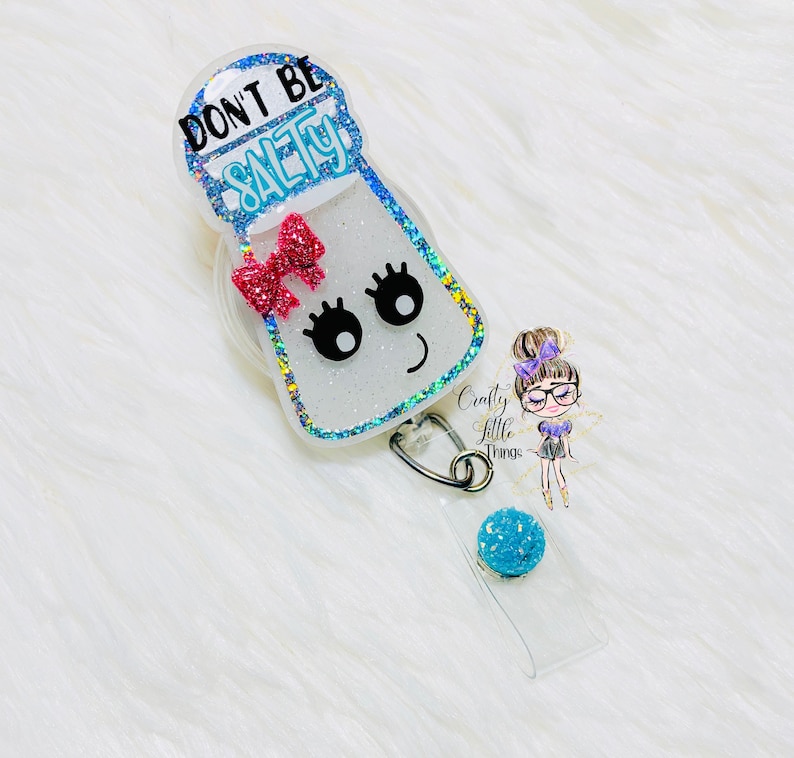 May include: A white salt shaker with glitter and a pink bow. The shaker has a smiling face with eyelashes and the text "Don't Be Salty" on the top. The shaker is attached to a clear badge holder with a blue glitter charm.