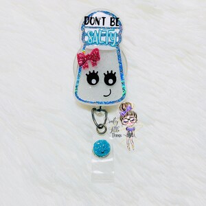 May include: A white glitter salt shaker with a pink bow and black eyelashes. The shaker has the text "Don't Be Salty" on it. The shaker is attached to a blue glitter retractable badge reel.