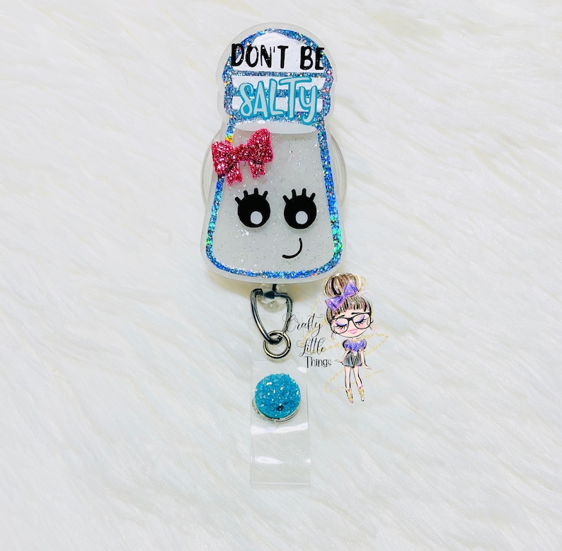 May include: A white glitter salt shaker with a pink bow and black eyelashes. The shaker has the text "Don't Be Salty" on it. The shaker is attached to a blue glitter retractable badge reel.