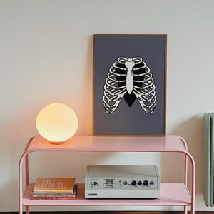 Art Print of a skeleton ribcage with a black heart encased onside on a blue/grey background shown displayed sat in a wooden frame in a mid-century modern home on a pink trolley against a white wall with green radiator.