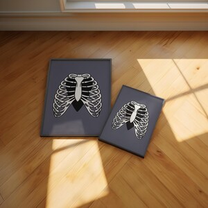 Art Print of a skeleton ribcage with a black heart encased onside on a blue/grey background shown laid out on the floor in A4 and 4 x 6 inch size black frames.