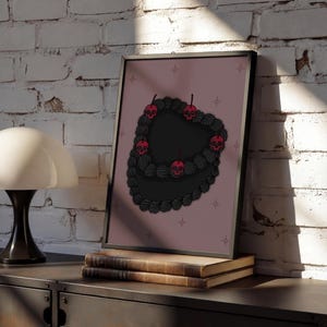 Könnte beinhalten: Gerahmter Kunstdruck mit einem schwarzen herzförmigen Kuchen-Design mit roten Totenkopf-Kirschen. Das Kunstwerk ist vor einem rosa Hintergrund mit Sternakzenten platziert. Der Druck wird auf einem dunklen Schrank mit Büchern ausgestellt.