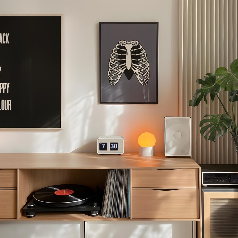 Art Print of a skeleton ribcage with a black heart encased onside on a blue/grey background shown in a black frame on the white wall of a mid-century style home with black record player and wooden sideboard.