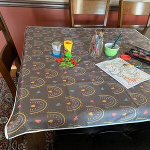 May include: A table covered in a dark gray tablecloth with a colorful rainbow and heart pattern. There are art supplies on the table, including paint, brushes, and a coloring page featuring two cartoon characters.