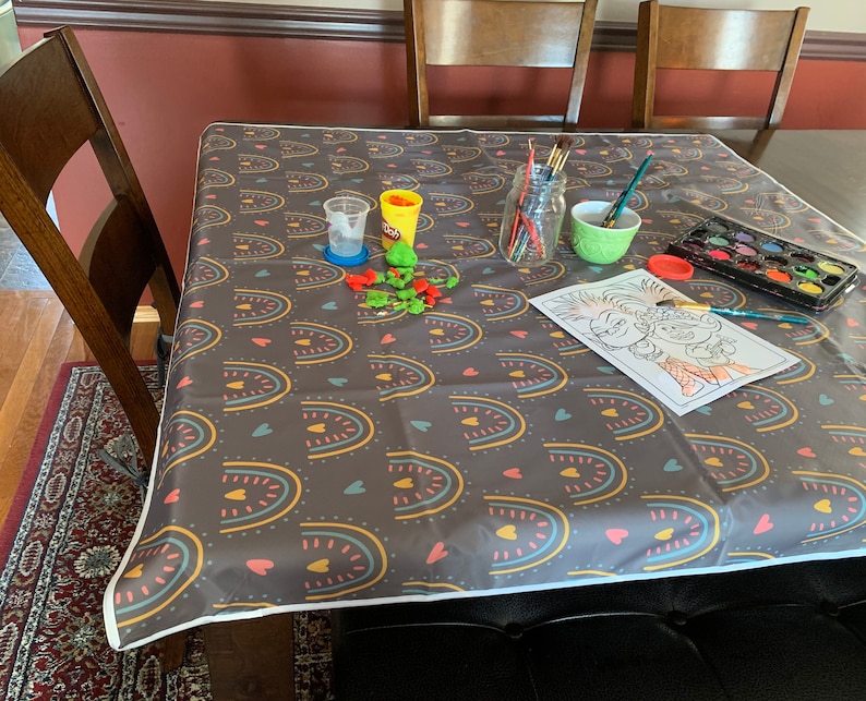 May include: A table covered in a dark gray tablecloth with a colorful rainbow and heart pattern. There are art supplies on the table, including paint, brushes, and a coloring page featuring two cartoon characters.