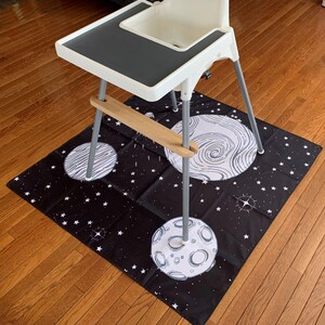 May include: A black and white space-themed play mat with three white moons and white stars on a black background. A white high chair with a gray tray is sitting on top of the mat.