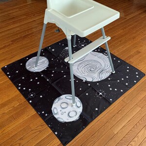 May include: A black and white space-themed playmat with planets and stars. A white high chair sits on top of the mat.