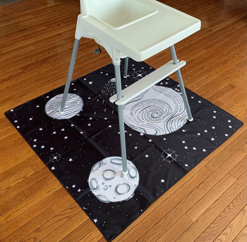 May include: A black and white space-themed playmat with planets and stars. A white high chair sits on top of the mat.