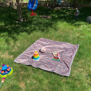 May include: A brown and white patterned picnic blanket with a rainbow design. A small ball, a stacking toy, and a small container are on the blanket.