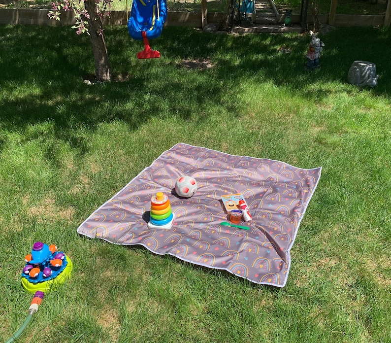 May include: A brown and white patterned picnic blanket with a rainbow design. A small ball, a stacking toy, and a small container are on the blanket.