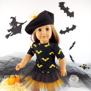 May include: A doll dressed in a Halloween costume. The outfit includes a black beret, a black t-shirt with yellow bat patterns, and a black and yellow tutu skirt. The doll is wearing white shoes. Halloween-themed decorations are in the background.