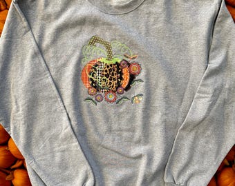 Op de afbeelding: Een grijze sweatshirt met een kleurrijke pompoenapplicatie. Het pompoenontwerp bevat luipaardprint, ruitjes en bloemmotieven. De achtergrond is gevuld met oranje pompoenen, wat een herfstthema creëert.