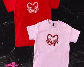 Könnte beinhalten: Zwei kurzärmelige T-Shirts, eines rot und eines rosa, werden gezeigt. Jedes Shirt hat ein herzförmiges Design, das aus zwei Skorpionen gebildet wird. Die Shirts sind aus weichem Material und für den Freizeitgebrauch geeignet.