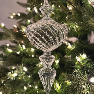 May include: A clear glass finial ornament featuring a bulbous top section with a swirling ribbed texture and a tapered, pointed bottom section. The ornament is suspended by a thin silver string and has a reflective, transparent surface.