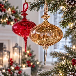 May include: Two teardrop-shaped glass ornaments hanging from a pine branch. One ornament is a deep, translucent red, and the other is a metallic gold. Both feature a ribbed, vertical texture, a rounded bulbous body, and a long, pointed finial tip.