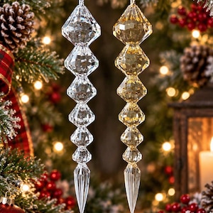 Pode incluir: Dois ornamentos pendentes em forma de pingente de gelo, um transparente e outro dourado, com uma série de segmentos facetados em forma de diamante que terminam numa ponta afiada. Ambos os ornamentos estão suspensos por fios prateados e dourados.