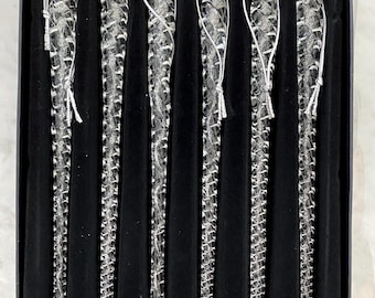 May include: A set of six clear glass icicle ornaments arranged in a black velvet-lined box. Each icicle features a twisted, spiral texture and includes a thin silver hanging cord attached to the top loop for display purposes.
