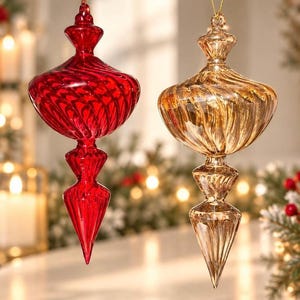 May include: Two finial-shaped glass ornaments hanging from gold strings. One ornament is a deep, translucent red, and the other is a translucent gold. Both feature a spiralling, ribbed texture that tapers to a pointed bottom and a decorative top.