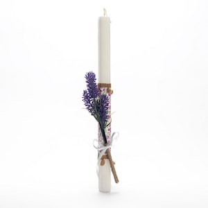 Puede incluir: Una vela blanca con un patrón floral y un ramo de flores moradas atado alrededor.
