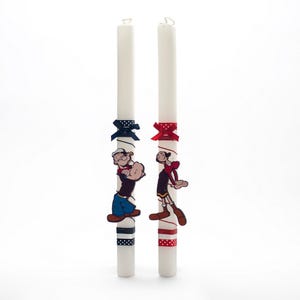 Puede incluir: Dos velas blancas decoradas con cinta roja y azul y personajes de dibujos animados. Los personajes son Popeye y Olivia.
