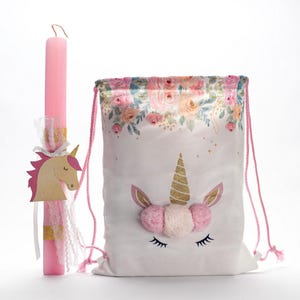 Puede incluir: Una bolsa de cordón rosa y blanca con un diseño de unicornio. La bolsa tiene un patrón floral en la parte superior y una cara de unicornio con pompones rosas en la parte delantera. Una vela rosa con una decoración de cabeza de unicornio de madera está al lado de la bolsa.