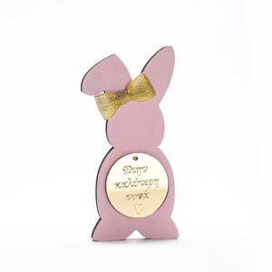 Puede incluir: Corte de conejo de madera rosa con un lazo dorado y una etiqueta redonda dorada que dice "Feliz Pascua" con un pequeño corazón.