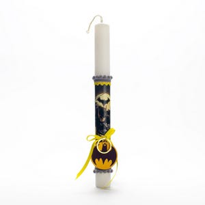 Puede incluir: Una vela blanca con ribete gris y amarillo. La vela tiene una imagen de Batman y una etiqueta amarilla con el logotipo de Batman.
