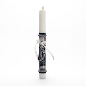 Puede incluir: Una vela blanca con un diseño estampado en gris y blanco. La vela tiene una cinta blanca atada alrededor y un pequeño amuleto plateado.