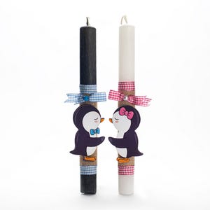 Puede incluir: Dos velas decorativas, una negra y otra blanca, adornadas con recortes de pingüinos. Las velas están envueltas con arpillera y cintas con estampados de cuadros vichy azules y rosas. Cada pingüino tiene una pajarita o lazo.
