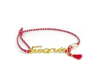 May include: A gold-colored bracelet with the Greek word "Γιορτή" in script. The bracelet features a red and white braided cord, a small pearl, and a red tassel. The design is simple and elegant.