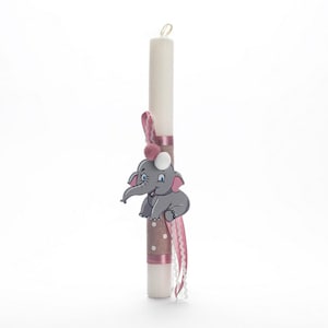 Puede incluir: Una vela blanca con una cinta rosa y marrón y una figurita de elefante gris. El elefante tiene orejas rosas y un lazo rosa.