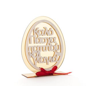 Puede incluir: Adorno decorativo en forma de huevo de Pascua con texto griego. El huevo dorado tiene letras blancas y una cinta roja. El adorno está diseñado para colocarse de pie.