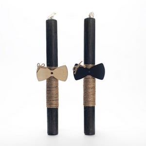 Puede incluir: Dos velas de Pascua negras decoradas con cuerda y pajaritas. Una pajarita es de madera y la otra es negra.