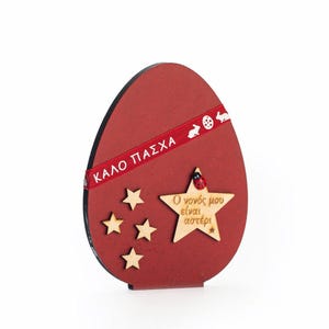 Puede incluir: Adorno de madera rojo en forma de huevo con una cinta roja que dice "KALO PAESXA" y una estrella de madera con el texto "O novos mou elvan aoteri" y una mariquita.