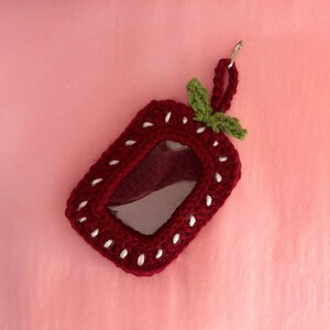 May include: A red crocheted strawberry-shaped ID badge holder with a clear plastic window and a green crocheted leaf on top.
