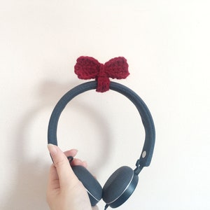 May include: Black headphones with a red crocheted bow on the headband. The headphones are held in a hand.