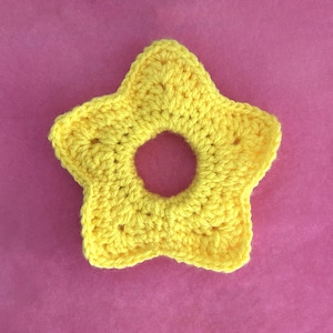 May include: A yellow crocheted star-shaped ring with a hole in the center. The ring is on a pink background.