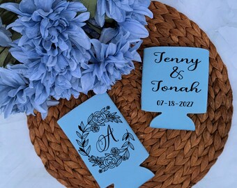 May include: Two light blue foam can coolers. One features a black floral wreath surrounding the letter A. The other displays the black text Jenny & Jonah and the date 07-18-2027. The items are positioned near a cluster of light blue flowers.
