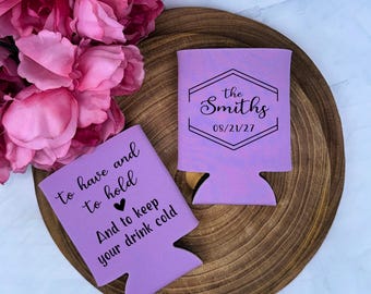 May include: Two lavender can coolers with black text. One cooler reads "To have and to hold, And to keep your drink cold." The other cooler has "the Smiths" and the date "08/21/27." The coolers are on a wooden tray with pink flowers.