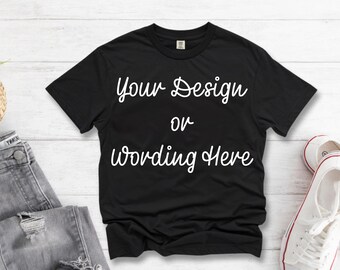 May include: Black t-shirt with the text "Your Design or Wording Here" in a white script font. The shirt is displayed on a white wooden surface, next to a pair of gray jeans and white sneakers.