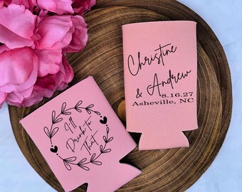 May include: Two light pink foam can coolers. One features a black floral wreath graphic with the text "I'll Drink to That". The other displays the black text "Christine & Andrew", the date "8.16.27", and the location "Asheville, NC" in a clean font.
