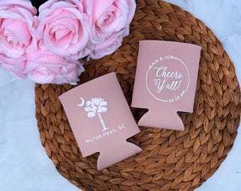 May include: Two pink can coolers with white text and graphics. One cooler features a palm tree and crescent moon, with "Hilton Head, SC" below. The other says "Jenn & Josh Cheers Y'all!" with a date. Pink roses are in the background.