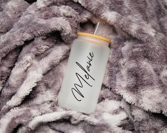 May include: A frosted glass tumbler with a bamboo lid and a reusable straw. The name "Melanie" is written in black script on the front of the tumbler. The tumbler is set against a soft, gray, plush blanket.