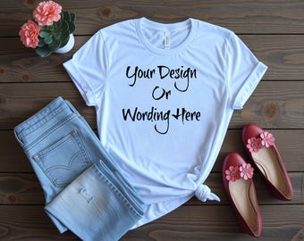 May include: A white t-shirt with the text "Your Design Or Wording Here" in black script, paired with light blue jeans and red flats adorned with pink flower accents. A small potted plant with pink flowers is also present.