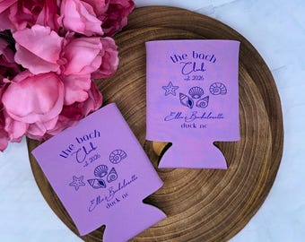 May include: Two light purple foam can coolers featuring dark purple text that reads the bach Club est 2026 Ellie's Bachelorette duck nc, accompanied by graphic illustrations of a starfish and three different seashells printed on the front.