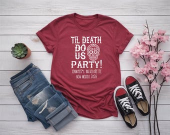May include: A maroon t-shirt with white text that reads "Til Death Do Us Party!" and a sugar skull graphic. The text below reads "Jennifer's Bachelorette New Mexico 2026". The shirt is paired with blue jeans and black sneakers.
