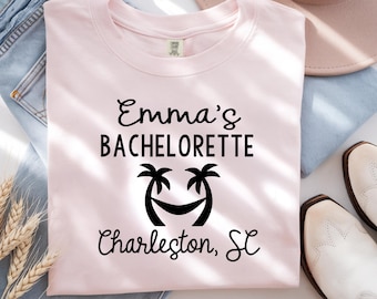May include: A light pink t-shirt with the text "Emma's Bachelorette Charleston, SC" in black script. The design includes a palm tree and hammock graphic. The shirt is laid on top of blue jeans, with a hat and white boots in the background.