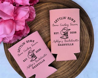 May include: Two light pink foam can coolers featuring black printed text that reads SETTLIN' DOWN, Same Cowboy Forever, EST. 2026, Ashlyn's Bachelorette, and NASHVILLE, accompanied by a graphic illustration of a cowboy hat resting on a cowboy boot.