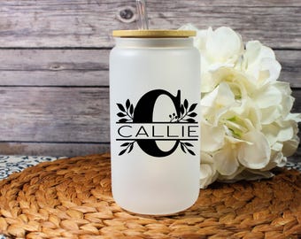 May include: A frosted glass tumbler with a bamboo lid and straw. The tumbler features a black monogram with the name "CALLIE" in a decorative font. The tumbler is sitting on a woven placemat.