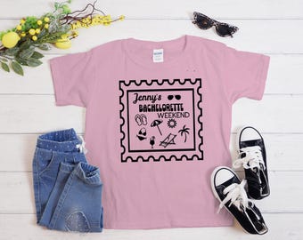 May include: A light pink t-shirt with the text "Jenny's Bachelorette Weekend" in a black stamp design. The design includes sunglasses, sandals, a cocktail, and a palm tree. A pair of blue jeans and black sneakers are also in the image.